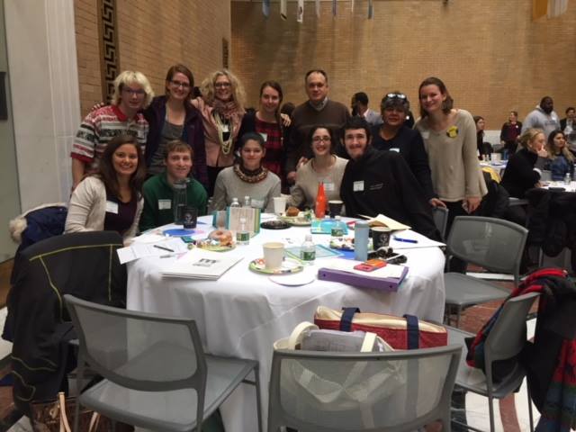 Group of PYD youth and staff standing behind a table with chairs in a wide, formal room