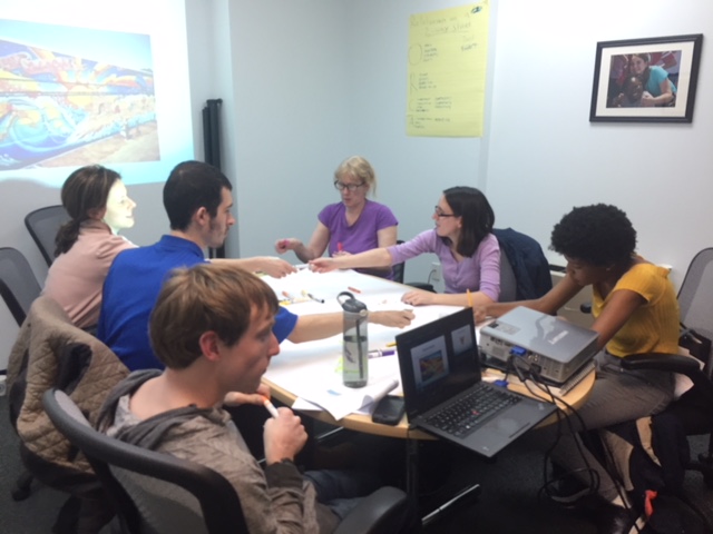 Group of young adults with various disabilities sitting around a table in a small conference room, with papers on the table in front of them and a picture being projected on the wall behind them.