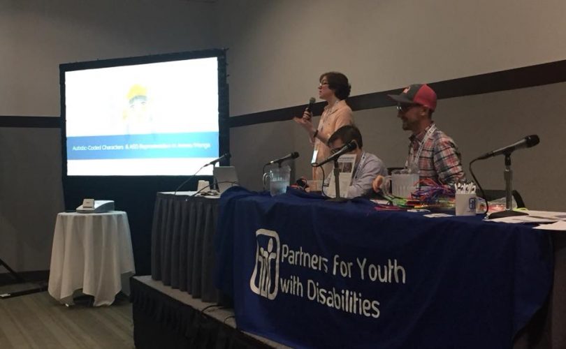 Jordan standing on a podium, introducing the panel presentation on the microphone, while her colleagues sit next to her.