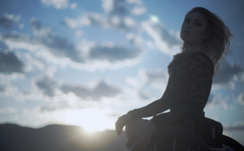 Young woman with long dirty blond hair in wheelchair and fitted black lace top, with low mountains and sun at the mountain line with blue sky and white clouds