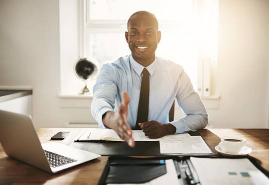 Black man in a shirt & tie reaching across a desk to shake the viewer's hand