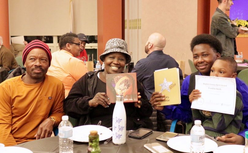 Photo of spirit award winner Franklin and three of his family members pose for a photo smiling and holding up Franklin's certificate as they sit down at a table