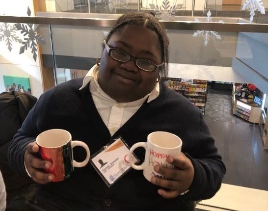 Donasia sits at a table at the Trade Show and holds 2 mugs that she made