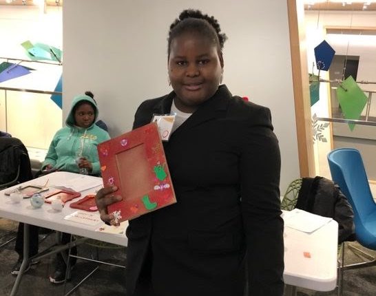 Sindy is wearing a black blazer as she smiles and holds up a red Christmas picture frame