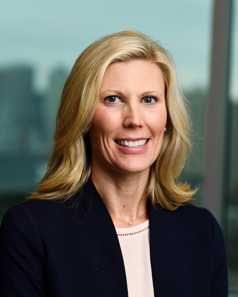 Bonnielin Swenor, a female with blonde hair wearing a navy suit jacket and light pink blouse, stands in front of a window overlooking a city scape.
