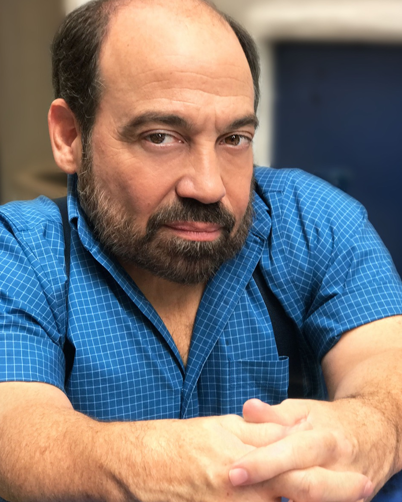Danny is a Little Person White male with a brown hair and beard and the hairline of a cruise ship captain. He seated at a bistro table outside, wearing a blue check shirt