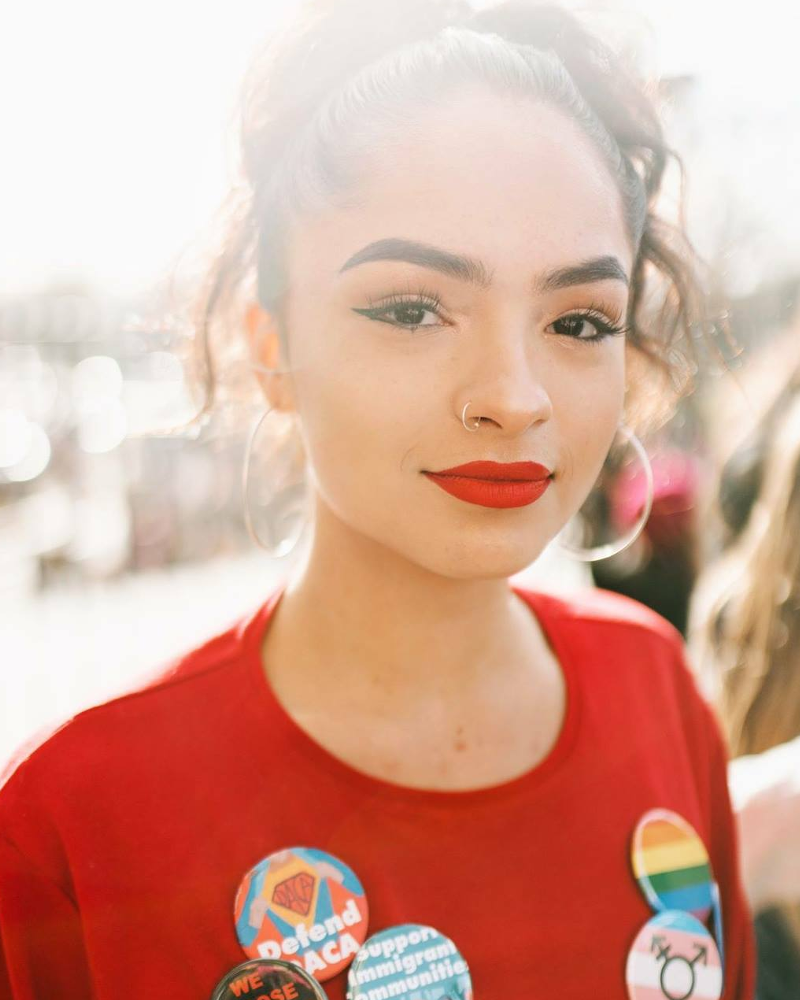 I am a light skin woman of color with black hair that is tied up in a curly ponytail. I am wearing red lipstick, mascara, black eyeliner and silver hoops. My shirt is plain red and I have 5 buttons on: one pink, white, and blue striped flag with the Trans* logo in the middle, one rainbow flag, one titled 