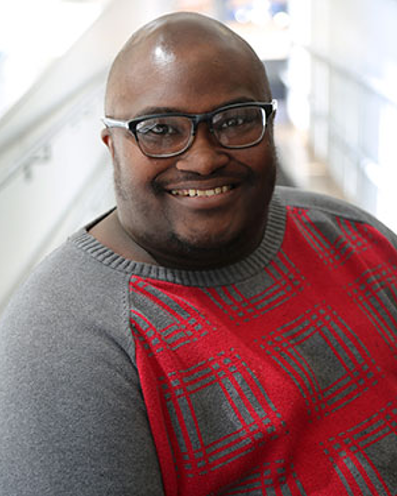 A bald Black man in his thirties. He is wearing glasses.
