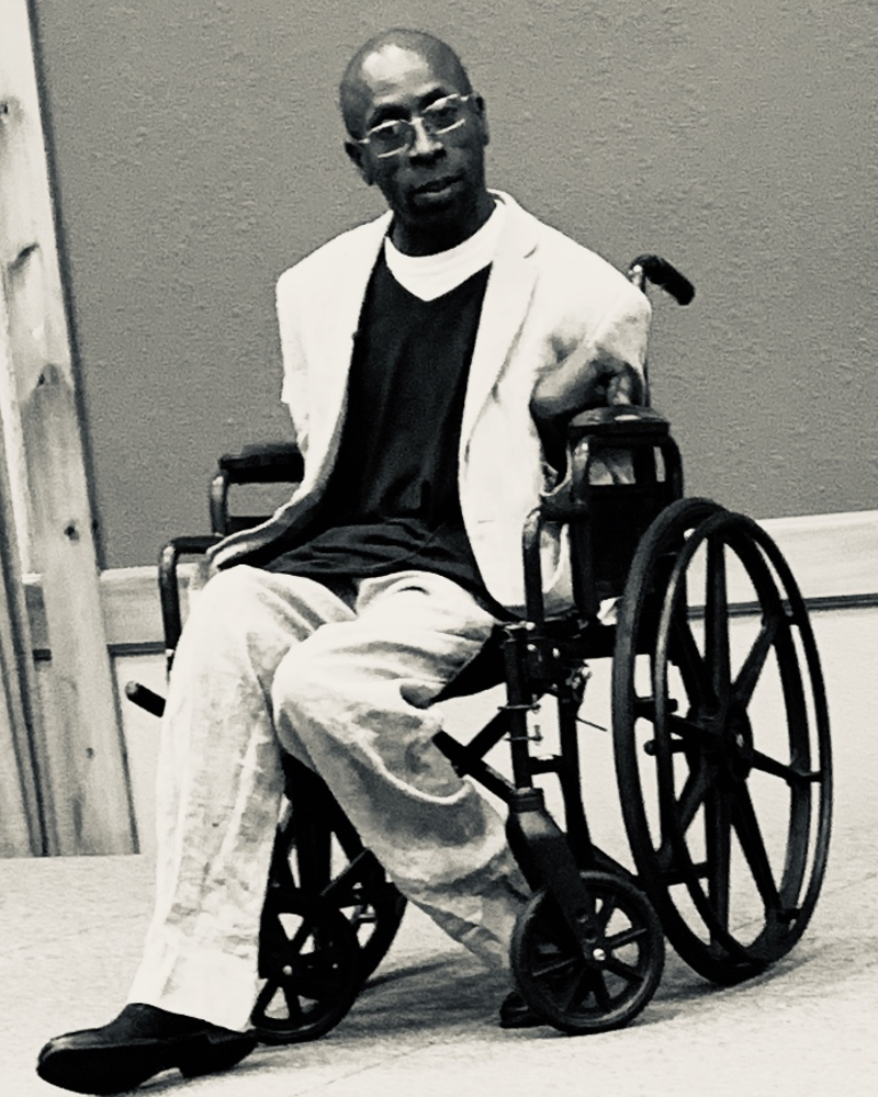 a photo of a black male state in a wheelchair on stage looking into the crowd while giving a speech