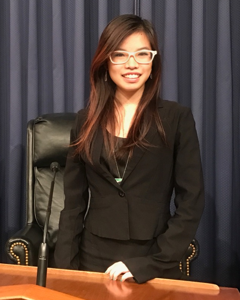 Kenna Chic is an East Asian woman with brown hair and white glasses. She is smiling at the camera and wearing a black blazer and business formal black dress. She is standing at a podium with a microphone in front of her. Behind her are a black armchair and blue curtains, and on the left side of the photo is the flag of the United States of America.
