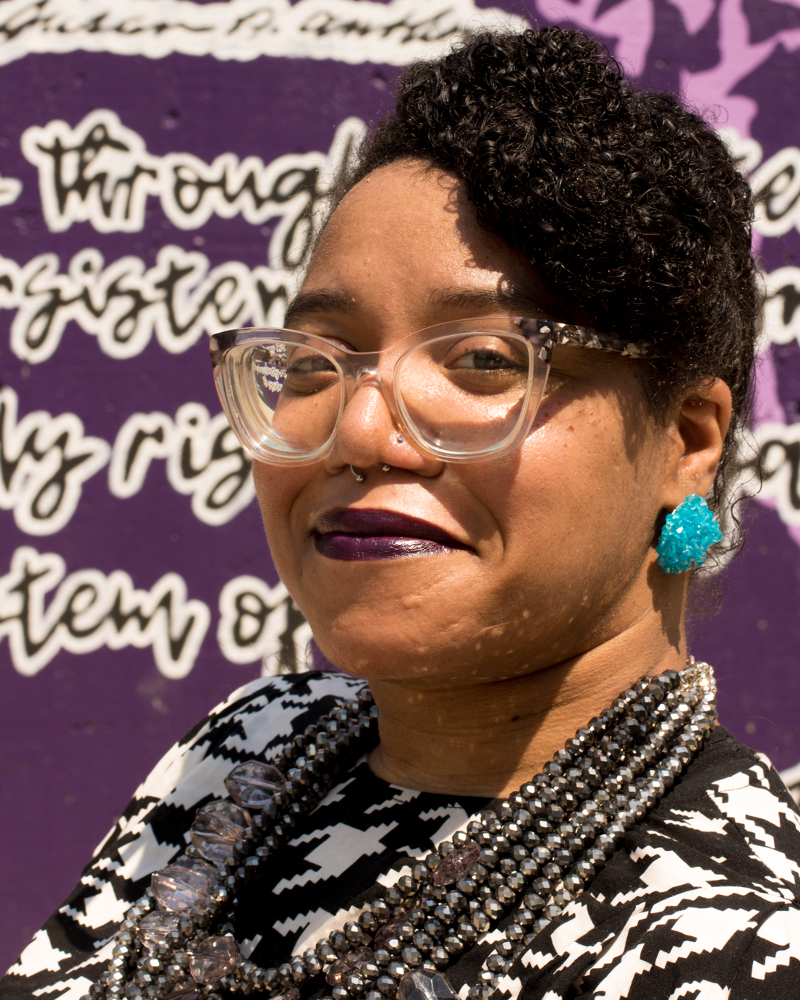 Picture of a honey skinned person sitting in a hot pink and black wheelchair with their hair braided into a swoop across their forehead. They are wearing a black and white houndstooth long sleeve shirt with fire hot pink pants, large clear glasses, Aqua colored earrings that look like rock candy, and a silver multistrand necklace. 