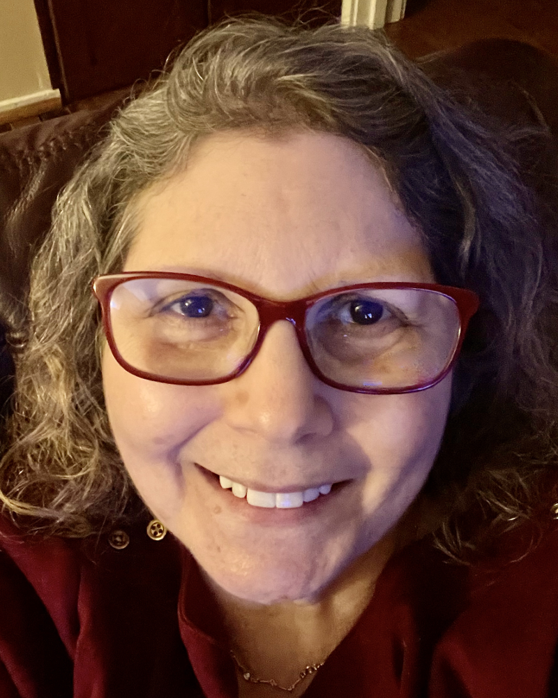 Older white woman with curly COVID-grey hair, red glasses and a matching sweater smiles for the camera.
