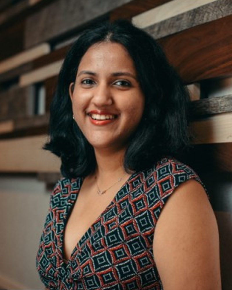 Meena, a brown woman with black short black hair smiling at the camera. She is wearing a blue-red square pattern dress and orange lipstick.