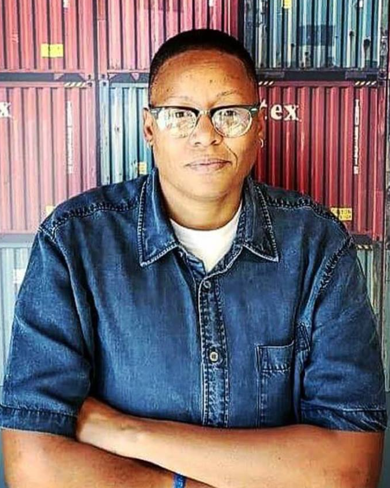 Brown skinned non-binary person with very short haircut, wearing browline glasses, blue button-down shirt. Najma's arm is crossed as they smile. In the background is a photo art of various ridged squares composed together. 