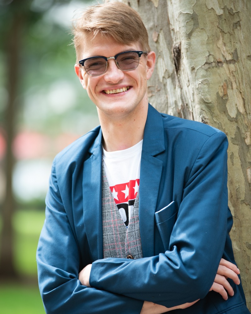 Photo of a blonde bespectacled man in a blue suit standing in front of a tree
