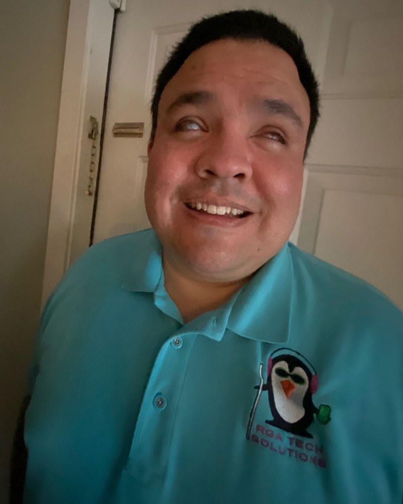 Raul has dark hair. He is wearing a blue company shirt with a logo of a penguin that says RGA Tech Solutions. The penguin is wearing headphones and is holding a phone and a cane.