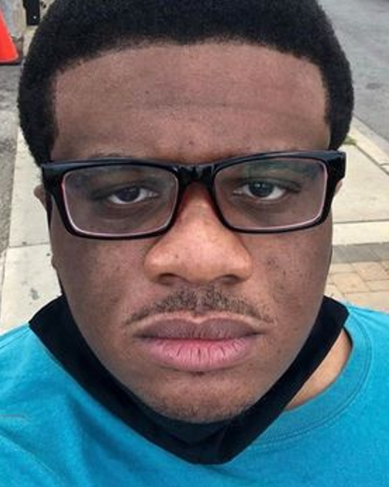 An African-American cis-male person, with medium complexion and mini black afro. He is wearing glasses, black mask around his neck, and a teal t-shirt