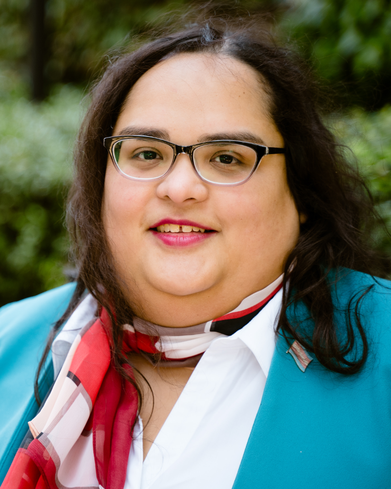 Latina woman in her early thirties. She is smiling towards the camera, wearing red lipstick, a blue blazer, white dress shirt and a red scarf. She has long wavy hair.