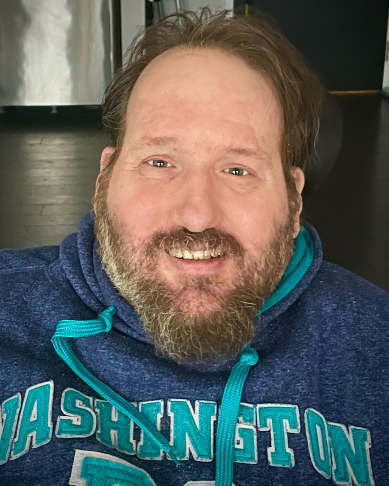 Middle aged White man with salt and pepper beard wearing blue hoodie, smiling with teeth.