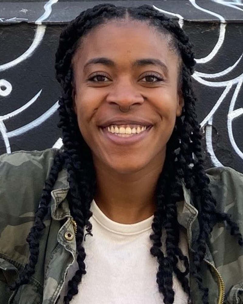 African American female smiling with black braids and nose ring. Wearing a white shirt and camo jacket. In front of a background that is black with white swirly lines.