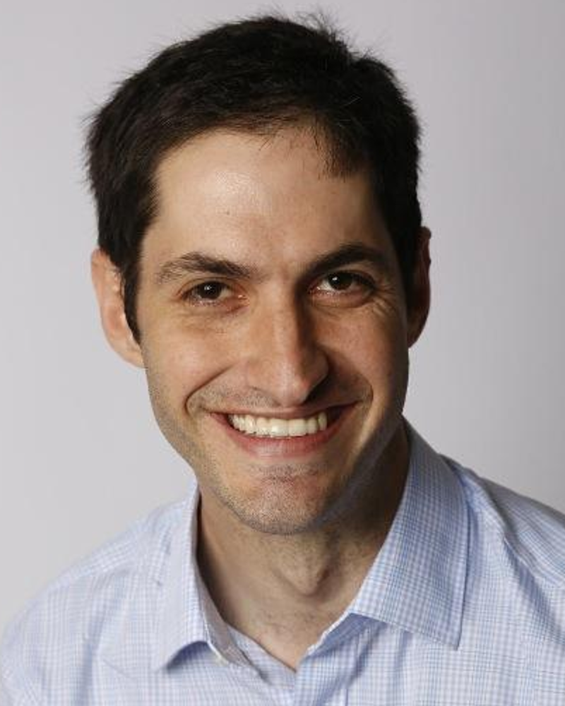 Eli has dark brown hair and dark brown eyes, a blue button down collared shirt, and he is smiling.