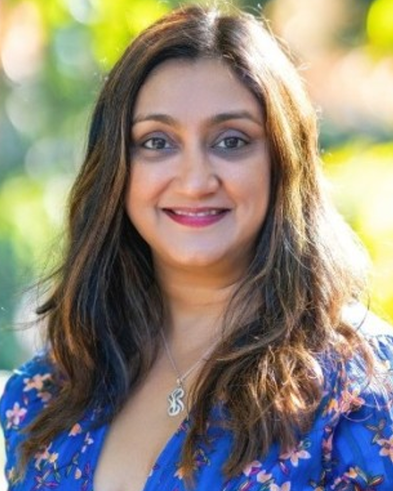 Gayatri is a woman of Indian origin with brown skin and long brown hair. She is wearing a blue dress with pink flowers on it. She is standing against a backdrop of green trees and sunlight.
