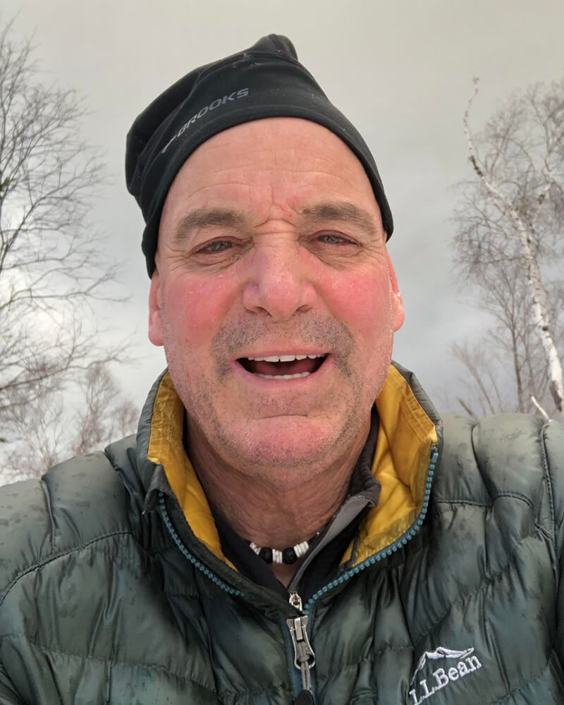 A white man wearing a black hat and green winter jacket is outside with trees in the background.