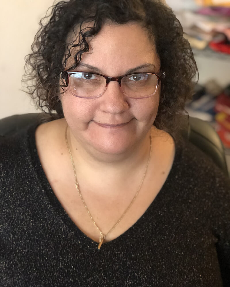 Mary, a Lebanese woman with short dark curly hair wearing glasses and a black V neck sweater. She is smiling at the camera.