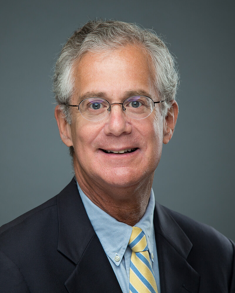 A white man wearing glasses, a light blue shirt, black blazer and yellow & blue stiped tie smiles