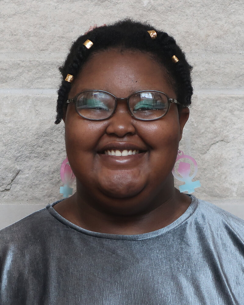 Oluwatobi is a smiling, Black person with their Afro pulled back. They are wearing a silvery blue blouse and are in front of a white wall.