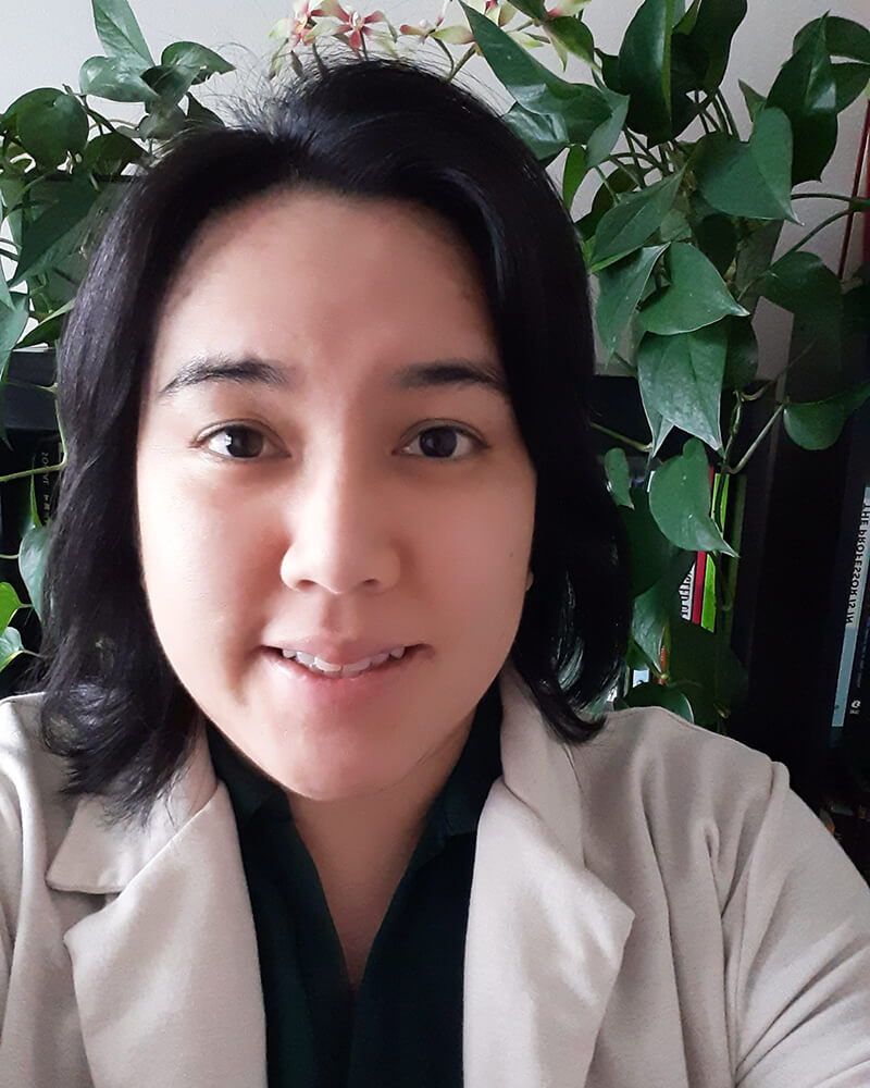 Queer crip Lucbanin Kapampangan Pilipinx who has light brown skin, dark brown eyes, and shoulder length black hair wearing a tan cardigan and dark green collar shirt smiling with dark green pothos plants in the background.