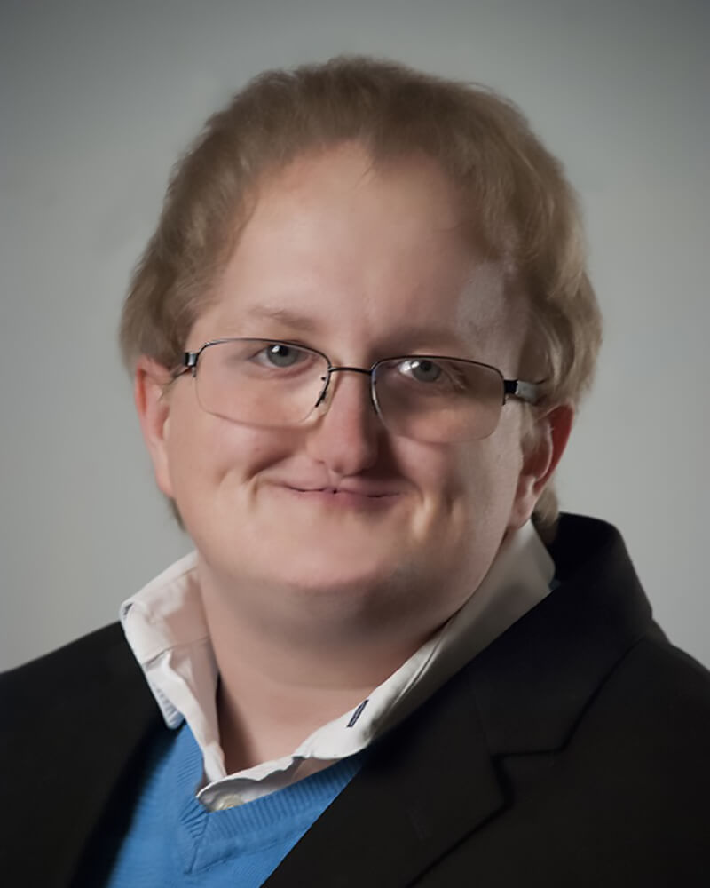 Photograph of a smiling white person with glasses, a cleft lip and palate, and dark blonde hair. They're wearing a black suit over a blue sweater and white undershirt.