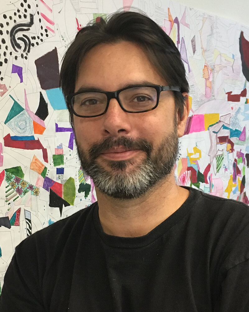 A man with a beard who is wearing dark glasses is standing along side a large colorful drawing he made at Painted Brain.