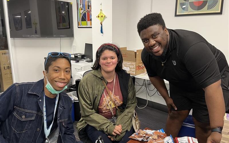 A participant in a denim jacket, a participant in a green jacket, and staff member Sayvion, wearing all black, smile while sitting in front of a table during game night.