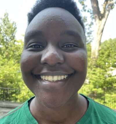 Ali is wearing a green shirt and smiling at the camera with nature in the background. She has short, curly black hair.