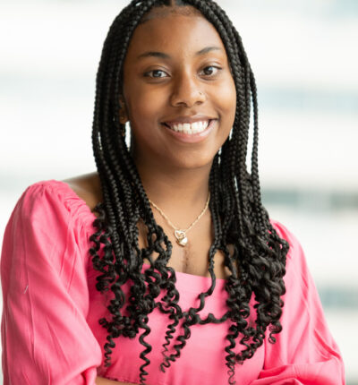 Amber has long dark brown hair in long braids with curly ends. Amber is wearing a pink blouse or dress and smiling.
