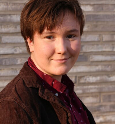 Emmett has short brown hair and is wearing a brown corduroy jacket with a red shirt underneath.
