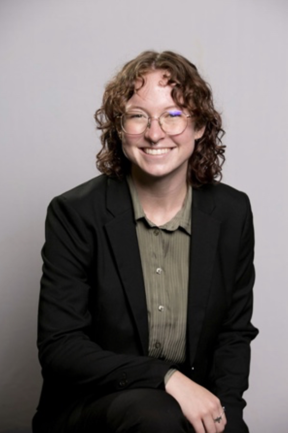 Natalie is wearing clear glasses and has shoulder length curly hair. They are wearing a sage green button up and a black blazer.