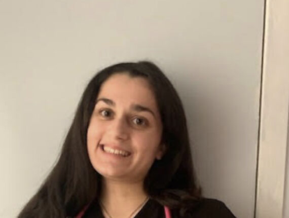 Mira stands against a white wall. She has long dark brown hair, brown eyes, and is smiling at the camera.