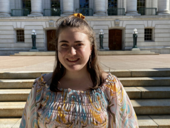Alexis, a white woman with shoulder-length brown hair, stands in front of a white building with large stone pillars. She is wearing a top that has swirls of the pastel-colored pink, yellow, blue, and white. 
