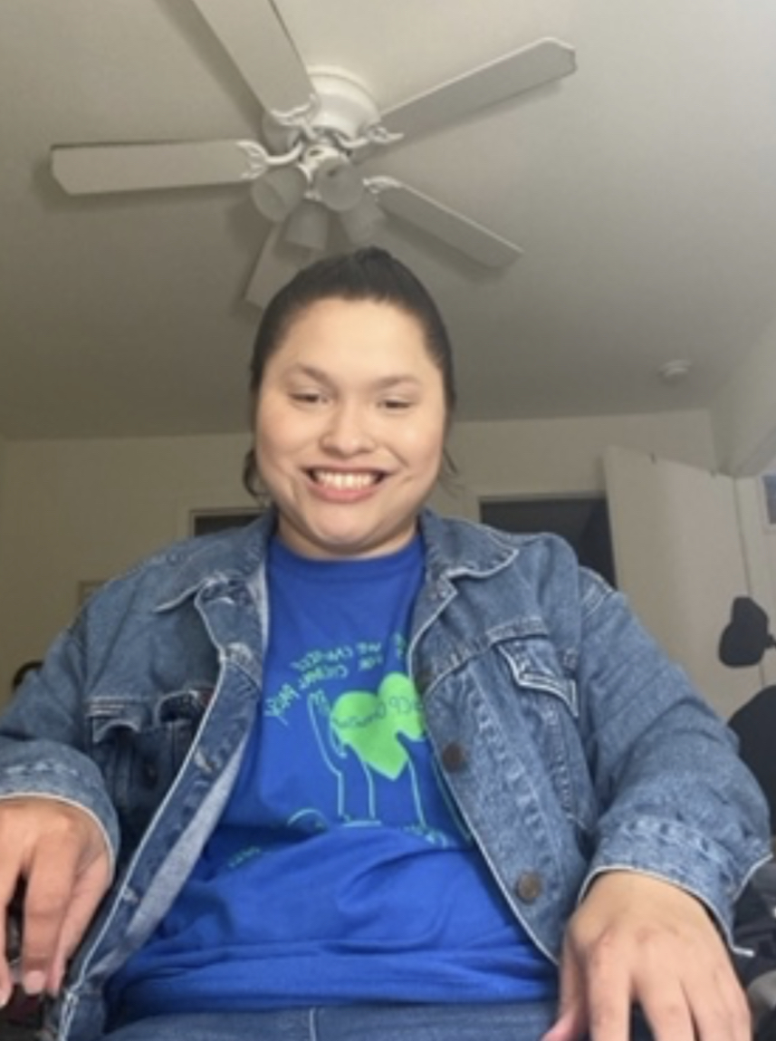 Alexa is sitting wearing a blue shirt and a jean jacket. She has brown hair in a ponytail or bun and is smiling.