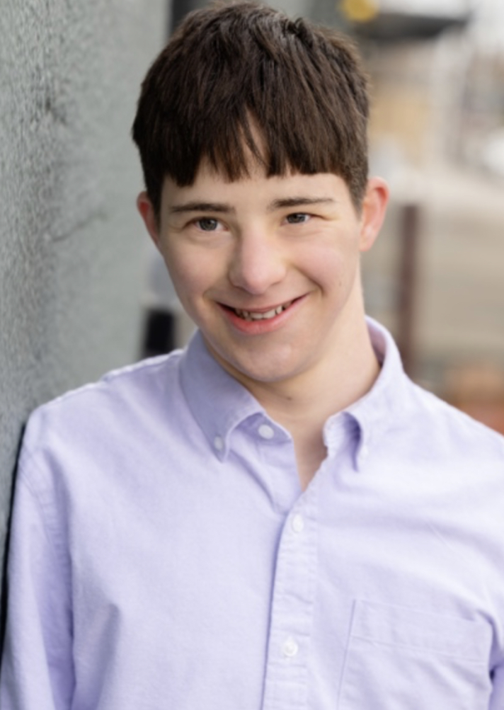 Dashiell is wearing a light blue button up shirt and smiling at the camera. He has short brown hair.