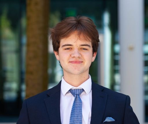 Nick has short brown hair. He is wearing a suit with a blue tie